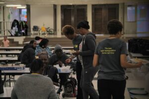 A group of people gathered in a room with long tables, engaging in conversation and sharing food. Some are seated, while others stand and serve food. Two people are wearing matching shirts with text on the back. The atmosphere appears friendly and casual.