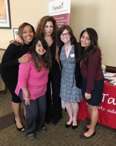 A group of five women smiling and posing for a photo in front of a display table with a red tablecloth featuring a logo and a freestanding banner. They are standing indoors, dressed in professional attire.
