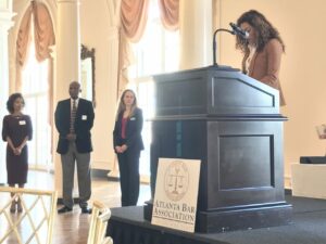 A woman stands at a podium with a microphone, speaking at an event. Three people stand in the background, listening. The podium displays a sign for the Atlanta Bar Association. The room is elegantly decorated with tall windows and draped curtains.