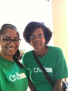 Two women smiling and standing side by side, wearing matching green T-shirts with white text. They are indoors, with a bright light in the background. Both have glasses and name badges.