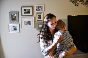 A woman with long dark hair holds a young child wearing a striped shirt. They are indoors with framed family photos on the wall and a TV nearby. The woman looks down gently at the child.