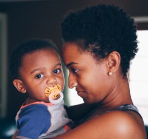 A woman holds a baby with a pacifier in her arms. They share a tender, close moment, both facing each other. The background is softly blurred, emphasizing their connection.