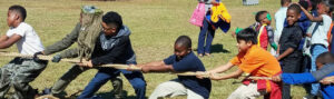 A group of children playing tug-of-war on a grassy field. They are pulling a thick rope with determination and teamwork. Some children in the background are watching or holding jackets and backpacks. The sun is shining brightly.