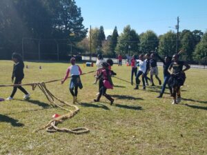 A group of children playing tug-of-war on a grassy field. They are pulling on a rope, and some are smiling and laughing. Cones and barrels are scattered in the background, and trees line the back of the field. It's a sunny day.