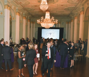 A formal gathering in an ornate ballroom with elegant chandeliers. Well-dressed individuals are socializing and holding drinks. A large screen in the background displays 