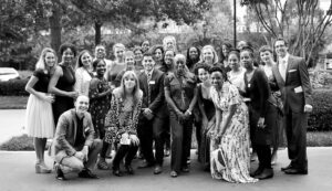 A diverse group of people stand together outdoors for a group photo, smiling and dressed formally. The setting is a park with trees in the background.