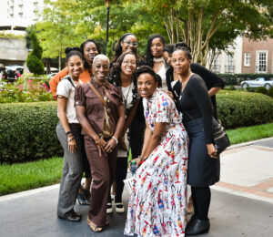 A group of nine women pose together, smiling outdoors in a garden setting. They are dressed in casual and formal attire, surrounded by greenery. Buildings and parked cars are visible in the background.