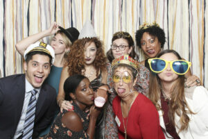 A group of people posing playfully with costumes and props in front of a colorful streamer backdrop. They wear hats, glasses, and hold funny items, smiling and making silly expressions.