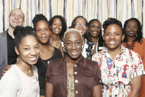 A diverse group of nine people, consisting of women and one man, stand closely together smiling. They are in front of a background with vertical ribbons in neutral colors. The atmosphere is joyful and friendly.