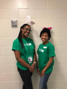 Two people wearing green shirts stand in front of a light-colored wall. One has bunny ears, and the other has a pink bow headband. They are smiling and standing close to each other.
