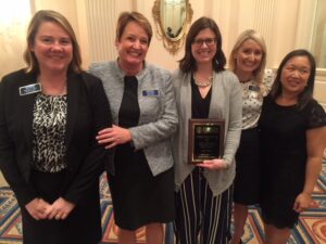 Five women are standing together indoors, smiling. One woman is holding a plaque. They are in professional attire, in a room with ornate décor and a carpeted floor.