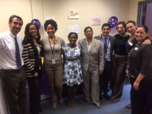 A group of nine people stands in a room with beige walls, smiling at the camera. The room has colorful drawings and purple balloons on the walls. Everyone is dressed in business casual attire.
