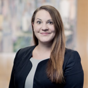 A person with long brown hair is wearing a black blazer over a gray top. They are standing indoors, smiling at the camera, with a blurred background.