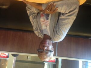 A person with short blonde hair and glasses is sitting at a table in a restaurant. They are wearing a gray jacket and a white t-shirt with red text. The restaurant has wooden panels and beverage signs in the background. The image is upside down.