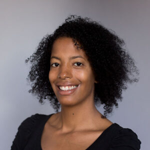 A person with curly black hair smiles at the camera. They are wearing a black top and the background is plain gray.
