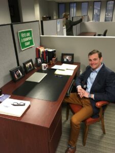 A man in business attire sits at an office desk with books, photos, and a laptop, smiling at the camera. A sign on the partition reads, 