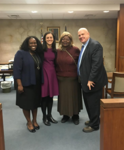 Four people stand together in a courtroom, smiling for a photo. Three women and one man are present, dressed in business attire. A flag and wood-paneled walls are visible in the background.