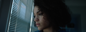 A person with curly hair looks pensively out a window with blinds, bathed in soft natural light.