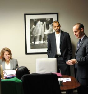 Three professionals having a discussion in an office. One woman is seated with documents, one man stands with hands in pockets, and another man stands speaking. A black and white photo hangs on the wall behind them.