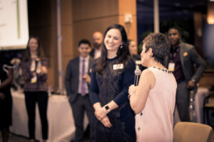A woman holding a microphone speaks to another woman who is smiling. They are in a room with several people in business attire, standing and observing. The setting appears formal with a screen and tables in the background.