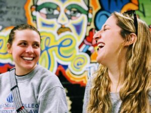 Two women laughing joyfully in front of a colorful graffiti wall. One wears a gray sweatshirt, while the other has long, wavy hair. Both are looking at each other, radiating happiness.