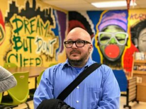 A man wearing glasses and a blue shirt stands in a room with vibrant graffiti on the walls, including the phrase 