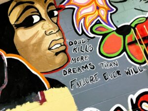 A vibrant mural features a woman's face with bold black eyeliner and red lips. Next to her, text reads, 