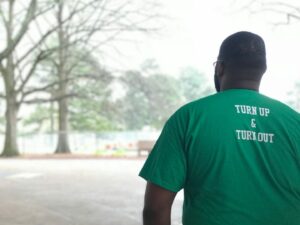 A person with short hair and glasses is wearing a green T-shirt with the slogan 