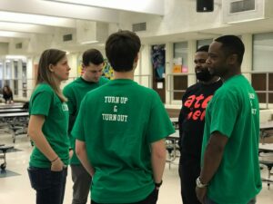 A group of five people stand in a school cafeteria, wearing matching green shirts with 
