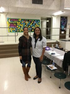 Two women standing together in a room with tables and chairs. One is wearing a plaid coat and brown boots, and the other is in a white patterned blouse and dark pants. A colorful bulletin board with notes is visible in the background.