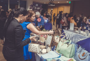 A group of people browsing a display of handbags and accessories on tables at an indoor market or event. The scene is busy, with various bags in different colors and styles prominently featured in the foreground.