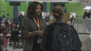 Two people are conversing in a busy indoor setting. One is facing the camera wearing a grey cardigan and lanyard, while the other has their back to the camera wearing a backpack. The background shows multiple people and green walls.