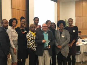 A group of people standing indoors, posing for a photo. One person at the center holds a trophy and smiles. They're in a conference room with a projector screen in the background. The group is diverse, with individuals dressed in business attire.