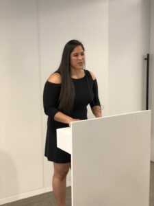 A woman with long hair stands at a white podium, speaking into a microphone. She is wearing a black dress and appears to be giving a presentation in a well-lit room.