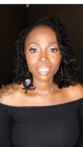 A person with shoulder-length curly hair is wearing a black off-the-shoulder top and hoop earrings. The background is dark, highlighting their warm smile and subtle makeup.