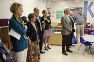A group of seven adults stand in a classroom, wearing business and casual attire. Some are holding papers. The room has children's furniture and educational posters on the walls.