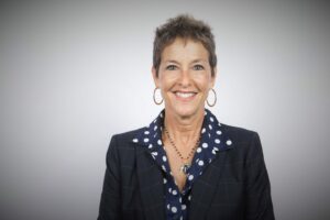 A woman with short hair smiles while wearing a polka dot shirt under a dark, checkered blazer. She has hoop earrings and a beaded necklace, posing against a plain, light background.