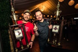 Two men smiling and holding large trophies with golden figures on top. They are standing under a dimly lit tent with hanging lights. One man is wearing a red shirt and cap, and the other is in a gray shirt gesturing with his index finger.