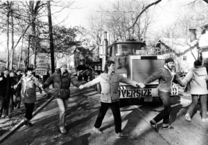 A black and white photo of protesters holding hands in front of a large truck on a residential street. One protester holds a sign saying, 