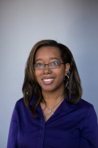 A person with long, brown hair is smiling at the camera, wearing glasses and a purple shirt. They also have a heart-shaped necklace and hoop earrings. The background is a plain, light gray.