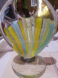 A multicolored glass award with abstract yellow, blue, and green patterns. The base is inscribed with text. A person is seated in the background.