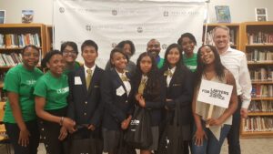 A group of students in school uniforms and adults in green shirts are smiling and standing together in a library. One person holds a sign that reads 