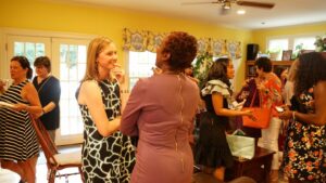 A group of women mingling and conversing in a brightly lit room with yellow walls. Some hold drinks and snacks. The atmosphere appears casual and friendly. Various women are standing and seated, engaging in conversation.