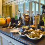 A bar counter with various drinks and mixers. Bottles and glasses are arranged neatly, with fresh ingredients like olives and celery in bowls. The background reflects a person working, and large windows let in natural light.