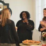 A group of five women are socializing in a well-lit room with a round table. Some hold drinks while one gestures. There are plates of muffins and flowers on the table. Large windows and a decorative mirror are in the background.
