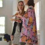 Two women standing indoors, engaging in conversation. One is holding a coffee cup, wearing a floral dress, and the other holds some papers, dressed in a patterned outfit. They appear to be in a well-lit room with large windows.