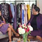 Two women in purple outfits are engaged in conversation, seated on chairs. Behind them, a clothing rack displays colorful garments. A glass vase with vibrant flowers sits on the table in the foreground.