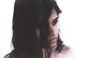 A woman with long dark hair looks to the side against a plain background. Her expression appears contemplative or somber, with her hair partially covering her face.