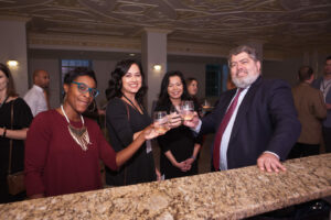 Four people are standing at a bar, smiling and holding drinks in a celebratory toast. The setting appears to be a formal social gathering or event, with several other attendees visible in the background.