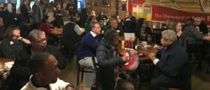 A diverse group of people sitting and standing inside a casual restaurant or bar. They are engaged in conversation, some looking towards the front of the room. Tables are scattered with food, drinks, and condiments. A sign is visible on the brick wall.